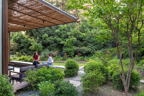A covered outdoor seating area with benches, surrounded by lush greenery and trees.