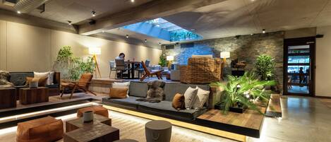 A modern lounge area with a sofa, chairs, and a dining table. There is a skylight above the seating area, and a person is working at a desk in the background.