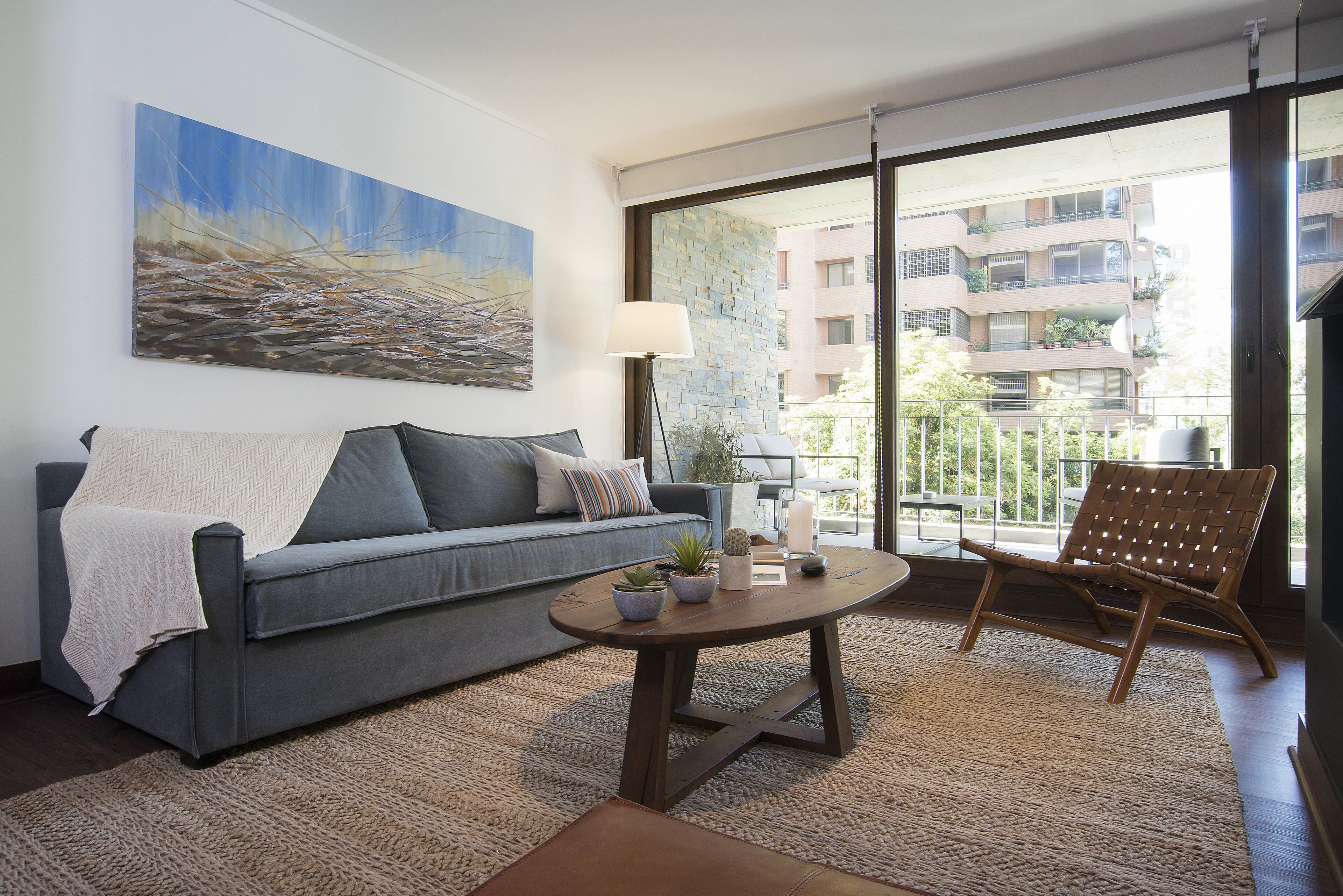 A modern living room with a grey sofa, a round coffee table, and a wicker chair. There is a large window with a view of a building and a balcony.