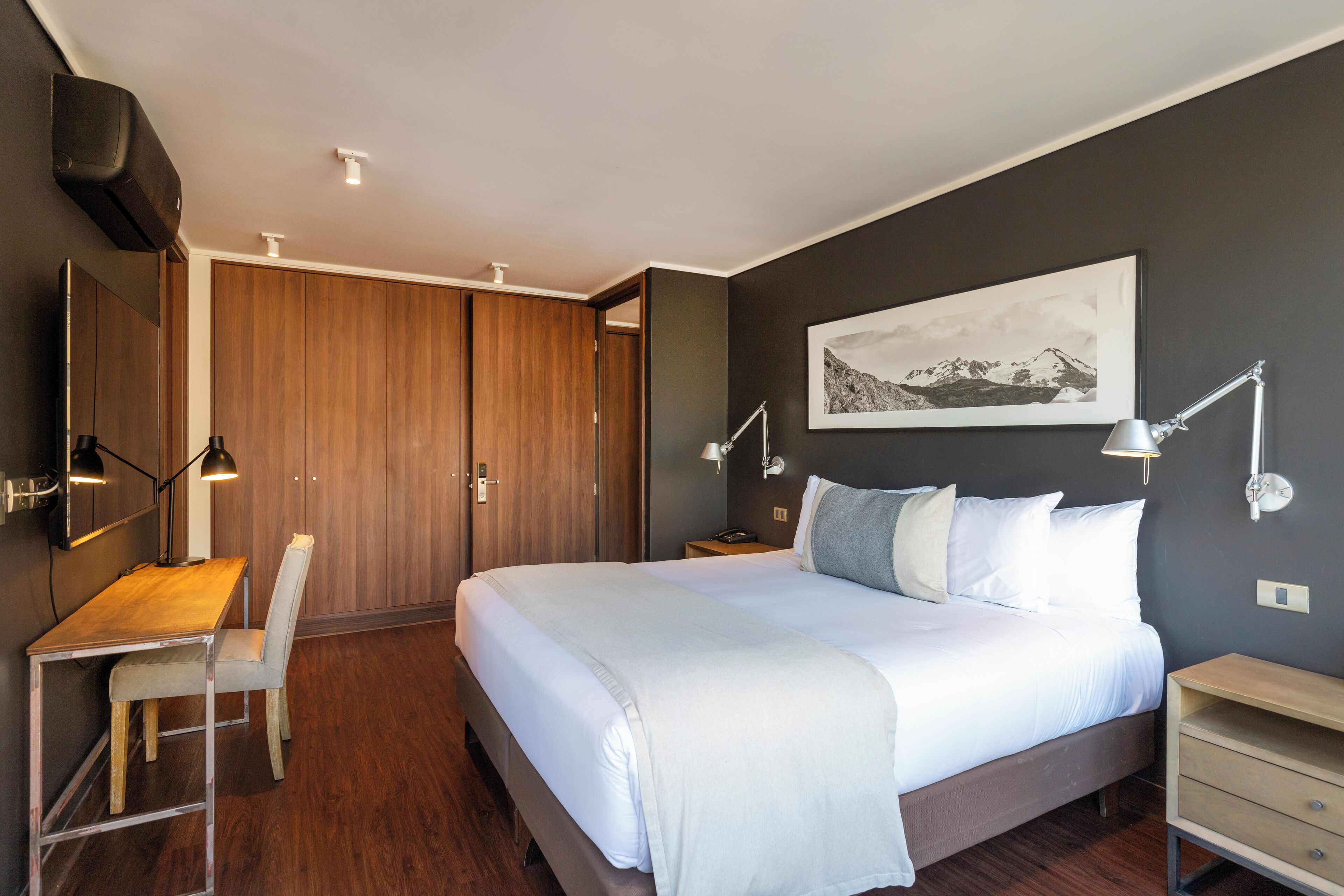 A modern bedroom with a large bed, a desk, and a chair. There is a wardrobe, a nightstand, and a wall-mounted lamp.