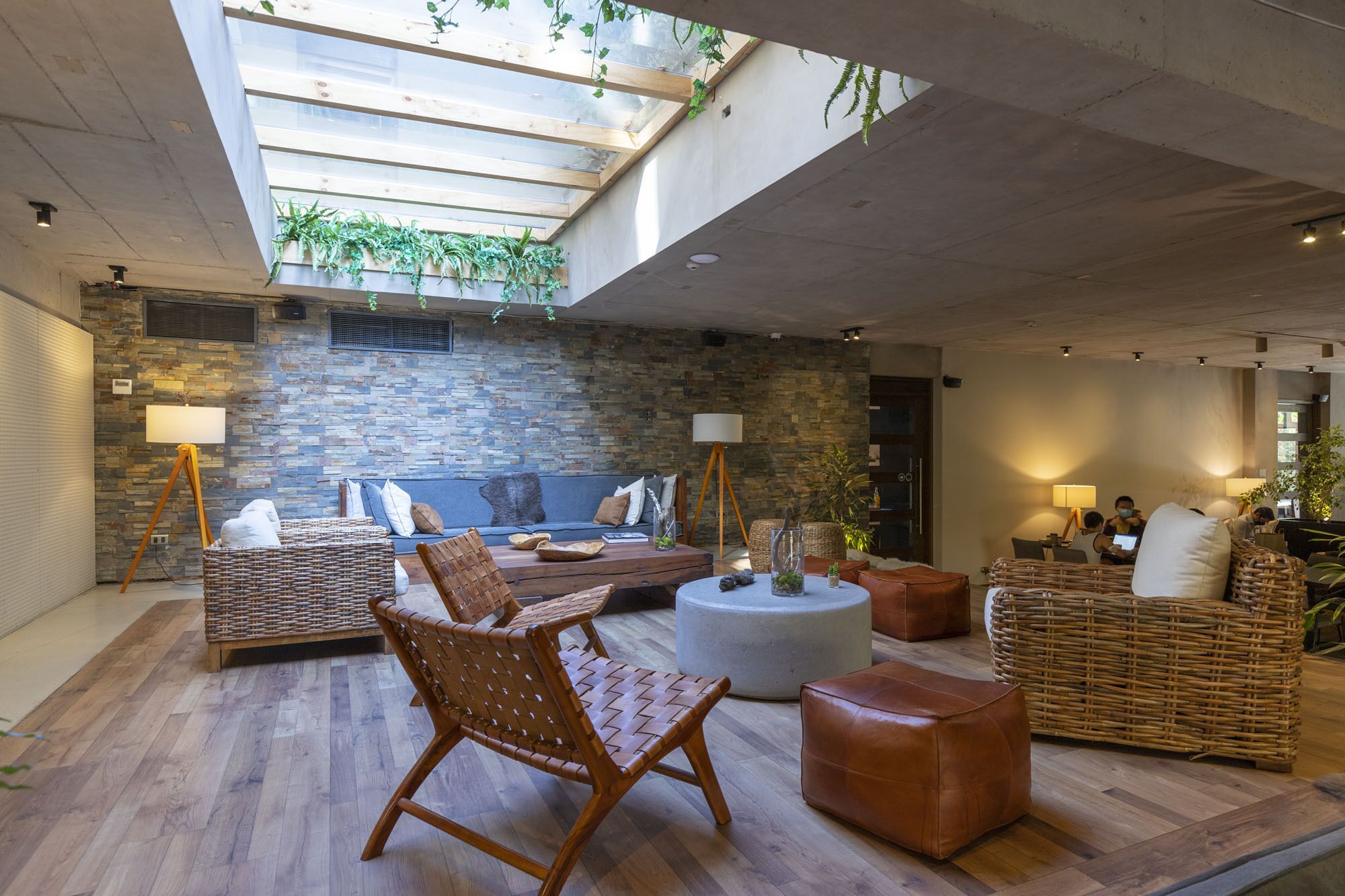 Un espacio interior amplio con pared de piedra, piso de madera y un techo alto con tragaluces. Hay muebles de mimbre, un reposapiés redondo y una zona de estar con cojines.