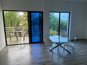 A spacious room with a glass table and chairs, large sliding glass doors leading to a balcony with a view of greenery and a building.