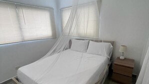 Une chambre à coucher avec un lit sous un dais, un ventilateur de plafond, une table de chevet avec une lampe et des fenêtres équipées de stores.