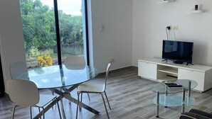Un salon moderne avec une table à manger en verre, deux chaises, une table basse en verre, un téléviseur à écran plat posé sur un meuble de télévision blanc et un ventilateur de plafond.