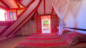 A bedroom with a large bed, a window with a red frame, and a view of the outside. - bungalows Cas'à Willy - Alamanda 3 (Sainte-Rose)
