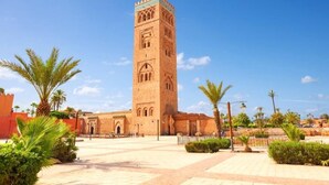 Una torre alta y ornamentada con detalles arquitectónicos intrincados, rodeada por una plaza empedrada con palmeras y un cielo azul despejado.