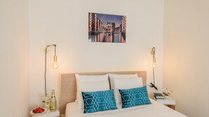 Une chambre d’hôtel bien rangée, avec un lit, deux tables de chevet, une petite table d’appoint, un tableau accroché au mur, et une petite table sur laquelle se trouvent une bouteille, un verre et une assiette de fraises.