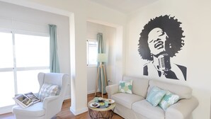 Un salon avec un fauteuil blanc, un canapé blanc, une table basse ronde sur laquelle se trouve un plateau de nourriture, et une fresque murale.