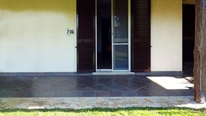 Entrada de un edificio con un porche adoquinado, una puerta corrediza de vidrio y contraventanas de madera.