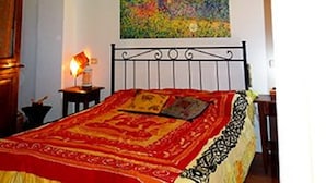 Une chambre avec un plafond en bois, un lit recouvert d’une couverture rouge et jaune, une table de chevet avec une lampe et un tableau accroché au mur.