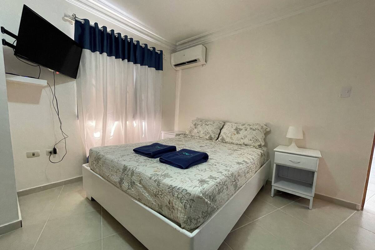 A bedroom with a bed, a TV mounted on the wall, an air conditioning unit, a nightstand, and a lamp.