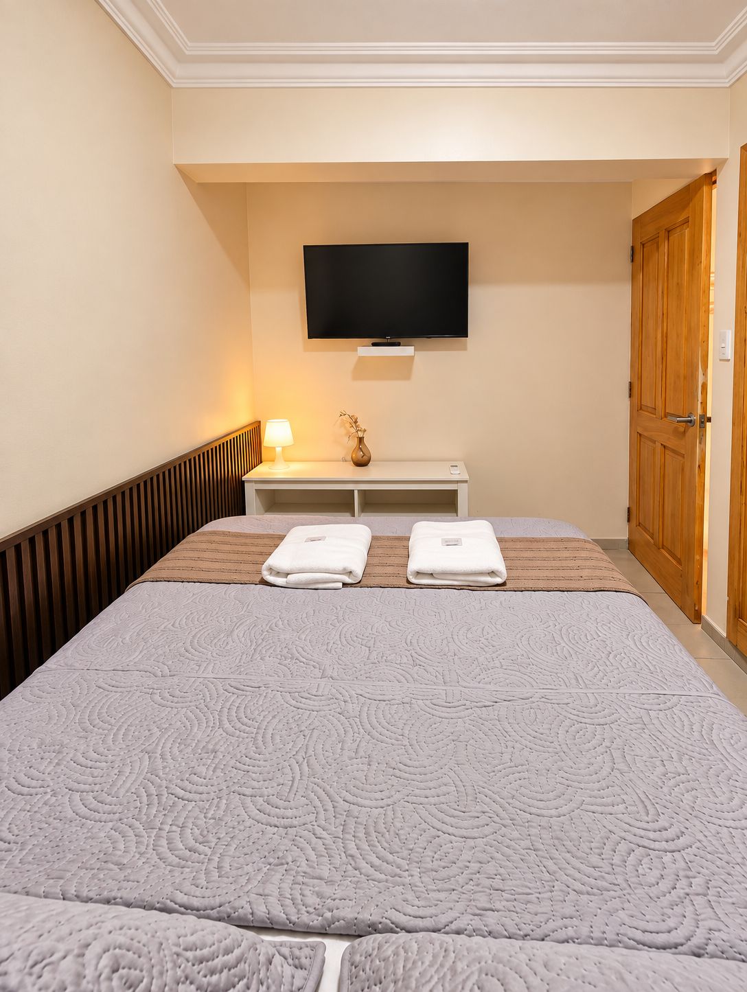 A hotel room with a bed, a flat-screen TV mounted on the wall, a bedside table with a lamp, and a wooden door.