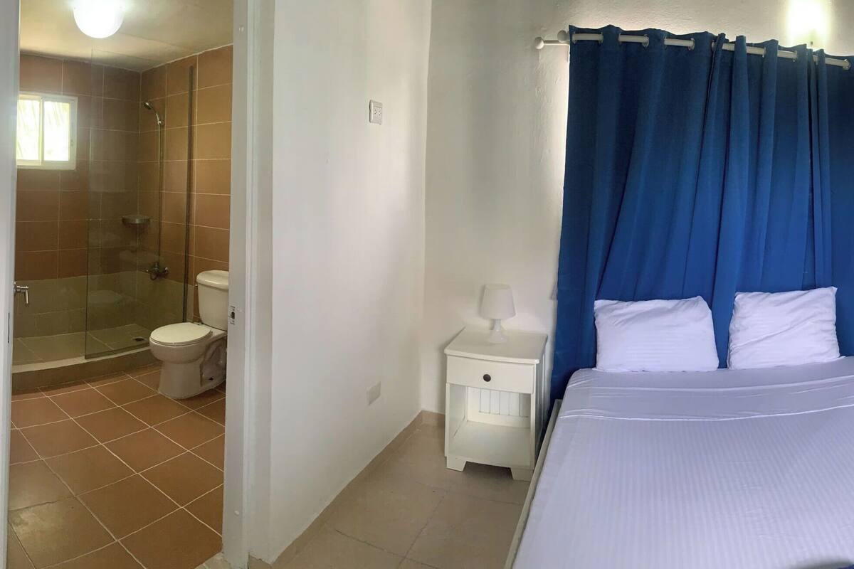 A small, compact room with a bed, a nightstand, a small table, a shower, and a toilet.