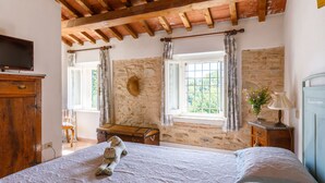 Una camera da letto rustica con parete in pietra, travi in legno, un letto con copripiumino bianco, un comò in legno e una finestra con tende.