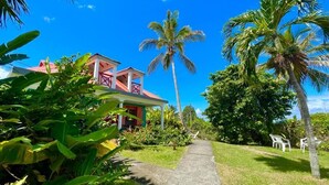Une maison avec un toit rouge et des murs blancs, entourée d’une végétation luxuriante et de palmiers.