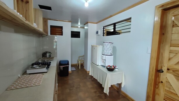 Un couloir étroit, avec une kitchenette sur la gauche, une table avec un distributeur d’eau et des bols sur la droite, et une porte en bois au fond.