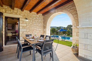 A stone-walled patio with a wooden table and chairs, a view of a swimming pool, and a covered area with a blue umbrella. - Villa Maza, beautiful stone villa in traditional village Maza, Chania (Maza)