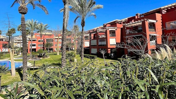 A hotel with red exterior walls, palm trees, and a clear blue sky. - Pool view private terrace ground floor Mijas Costa with barbecue by Solrentspain (Mijas Costa)