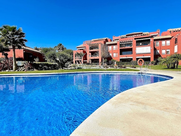 A swimming pool with a clear blue water surface, surrounded by a concrete deck. In the background, there are red buildings with multiple balconies and a palm tree.