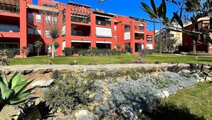 A red building with balconies, a garden area with plants, and a clear blue sky. - Pool view private terrace ground floor Mijas Costa with barbecue by Solrentspain (Mijas Costa)