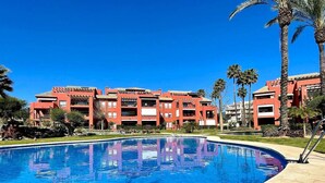 A swimming pool with a clear blue water surface, surrounded by a building with a red facade and multiple balconies. - Pool view private terrace ground floor Mijas Costa with barbecue by Solrentspain (Mijas Costa)