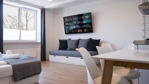 Ein modernes Wohnzimmer mit einer Couch, einem an der Wand montierten Fernseher, einem Esstisch und einem Fenster mit Jalousien.