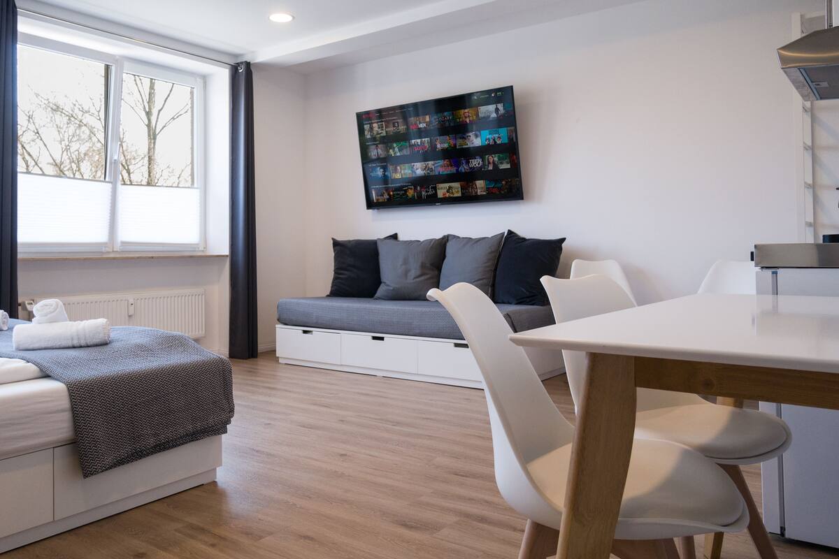 Ein modernes Wohnzimmer mit einer Couch, einem an der Wand montierten Fernseher, einem Esstisch und einem Fenster mit Jalousien.