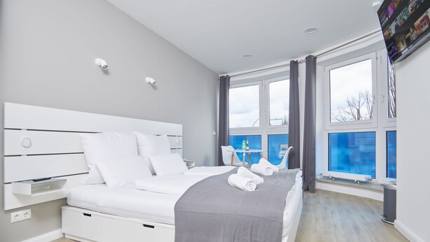 Ein modernes Schlafzimmer mit einem großen Bett, einem Nachttisch, einem Fenster mit Vorhängen und Blick ins Freie.