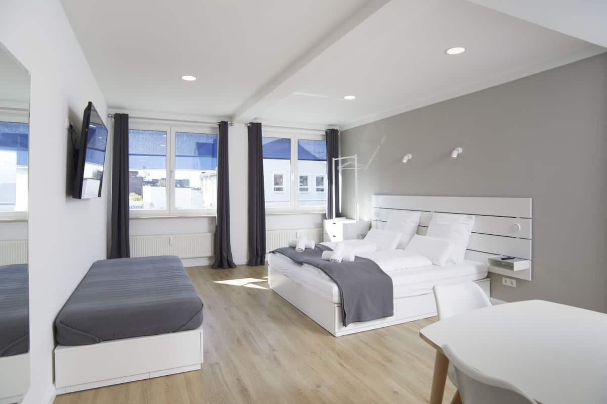 Ein modernes Hotelzimmer mit einem Bett, einem Schreibtisch und einem Stuhl. Es gibt Fenster mit Vorhängen, einen an der Wand befestigten Flachbildfernseher und einen kleinen Nachttisch mit einer Lampe.