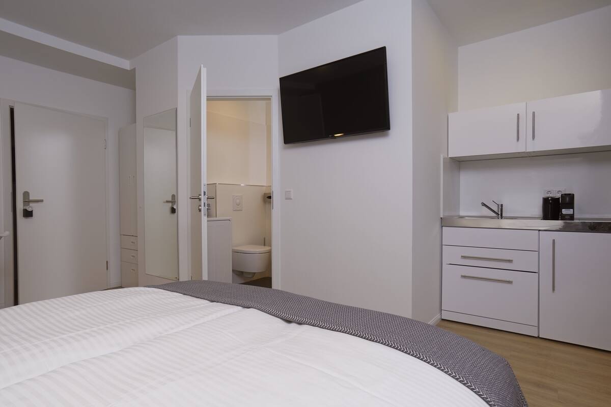 Ein Schlafzimmer mit Bett, einem an der Wand montierten Fernseher, einem Badezimmer mit Toilette und Waschbecken sowie einer Küchenzeile mit Schränken und einer Kaffeemaschine.