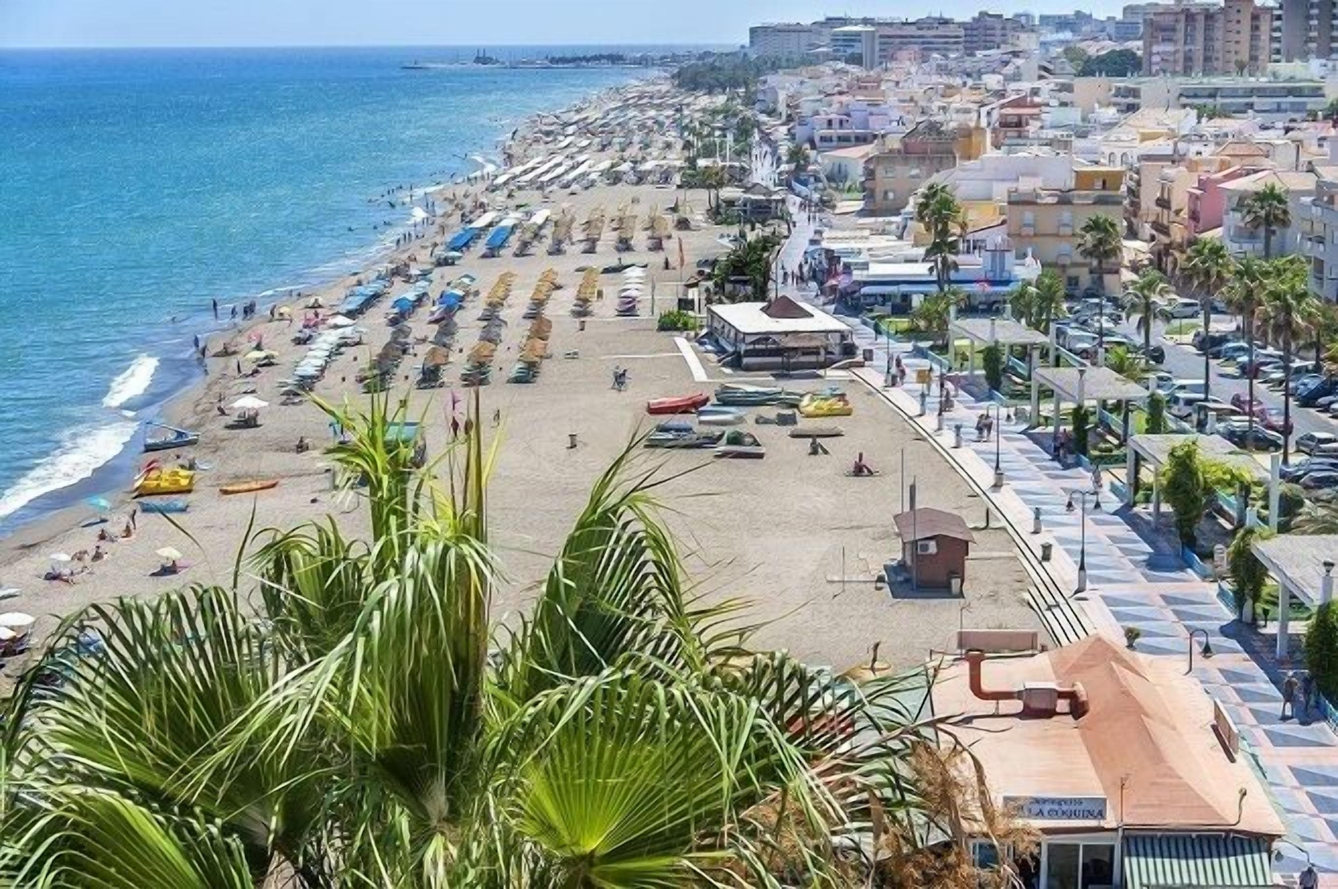 A beach with rows of umbrellas and sun loungers, a building with a sign, and a coastal cityscape in the background.