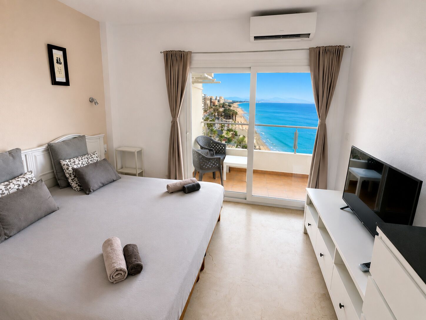 A hotel room with a bed, a TV, a balcony with a view of the sea, and a small table with chairs.