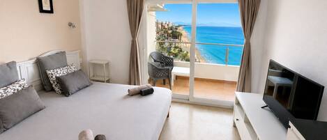 A hotel room with a bed, a TV, a balcony with a view of the sea, and a small table with chairs.