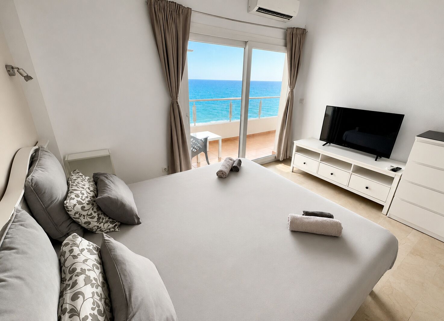 A hotel room with a large bed, a TV, and a balcony with a view of the sea.
