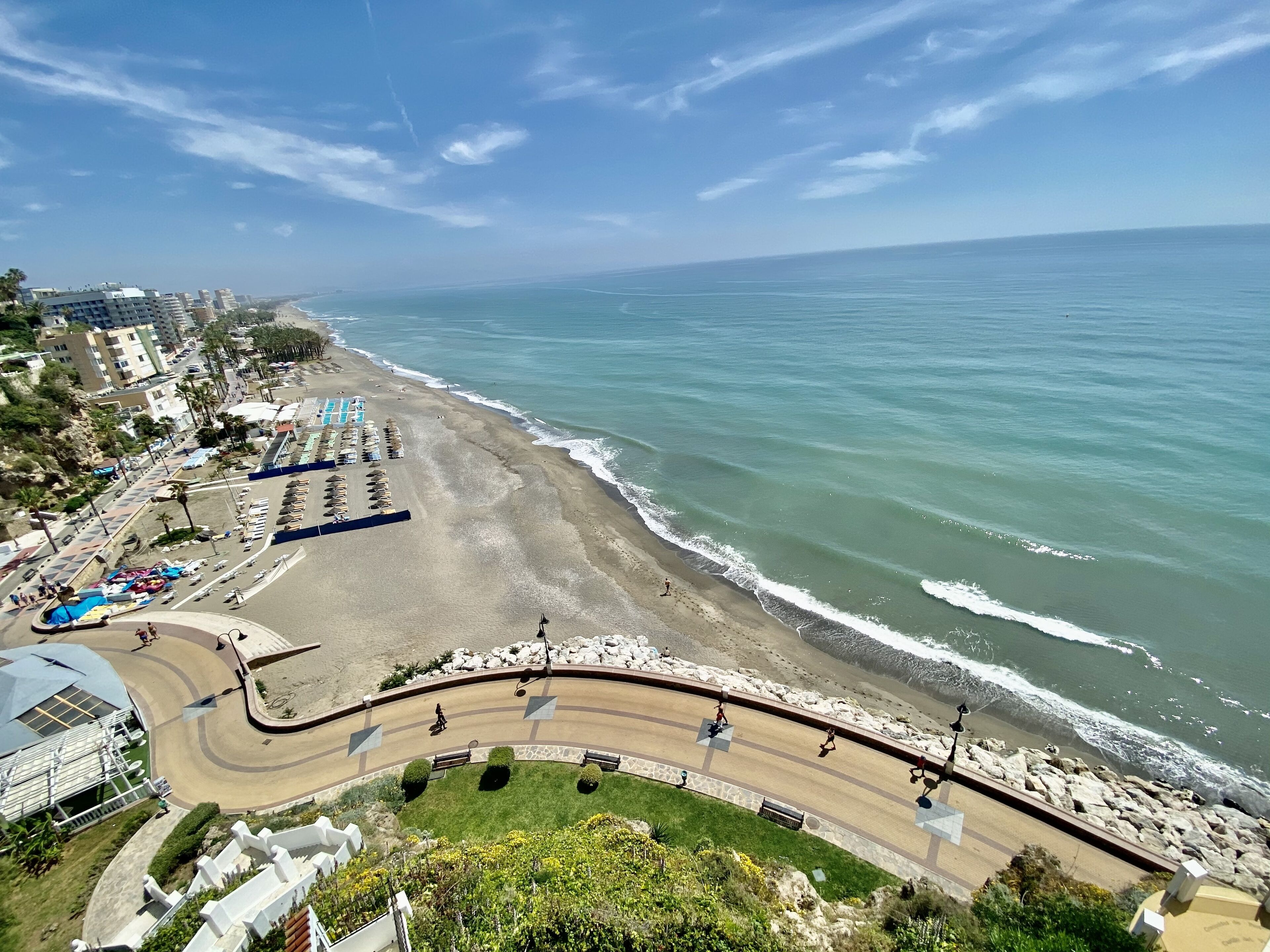 A coastal road with a beach on one side, a parking area, and buildings in the background.