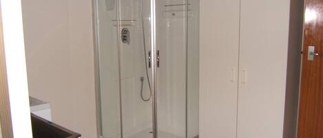 Cabina de ducha en esquina con puerta de cristal, armario blanco y un cubo verde encima.