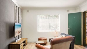 Un salon avec une table basse en bois, un fauteuil en cuir, un téléviseur fixé au mur et une fenêtre avec des stores.