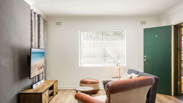 Un salon avec une table basse en bois, un fauteuil en cuir, un téléviseur fixé au mur et une fenêtre avec des stores.