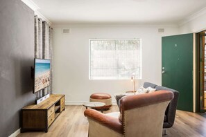 A living room with a wooden coffee table, a leather armchair, a television on the wall, and a window with blinds.