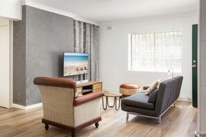 A living room with a television, a leather armchair, a grey sofa, a small wooden table, and a window with blinds.