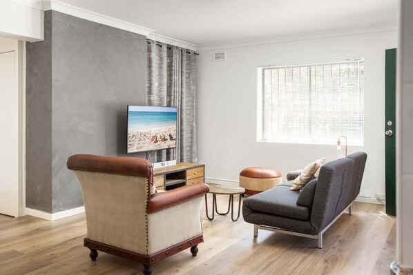 A living room with a television, a leather armchair, a grey sofa, a small wooden table, and a window with blinds.