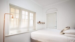 Une chambre à coucher avec un lit, une fenêtre avec des stores, une table de chevet avec une horloge et un miroir.