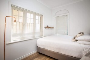 A bedroom with a bed, a window with blinds, a bedside table with a clock, and a mirror.
