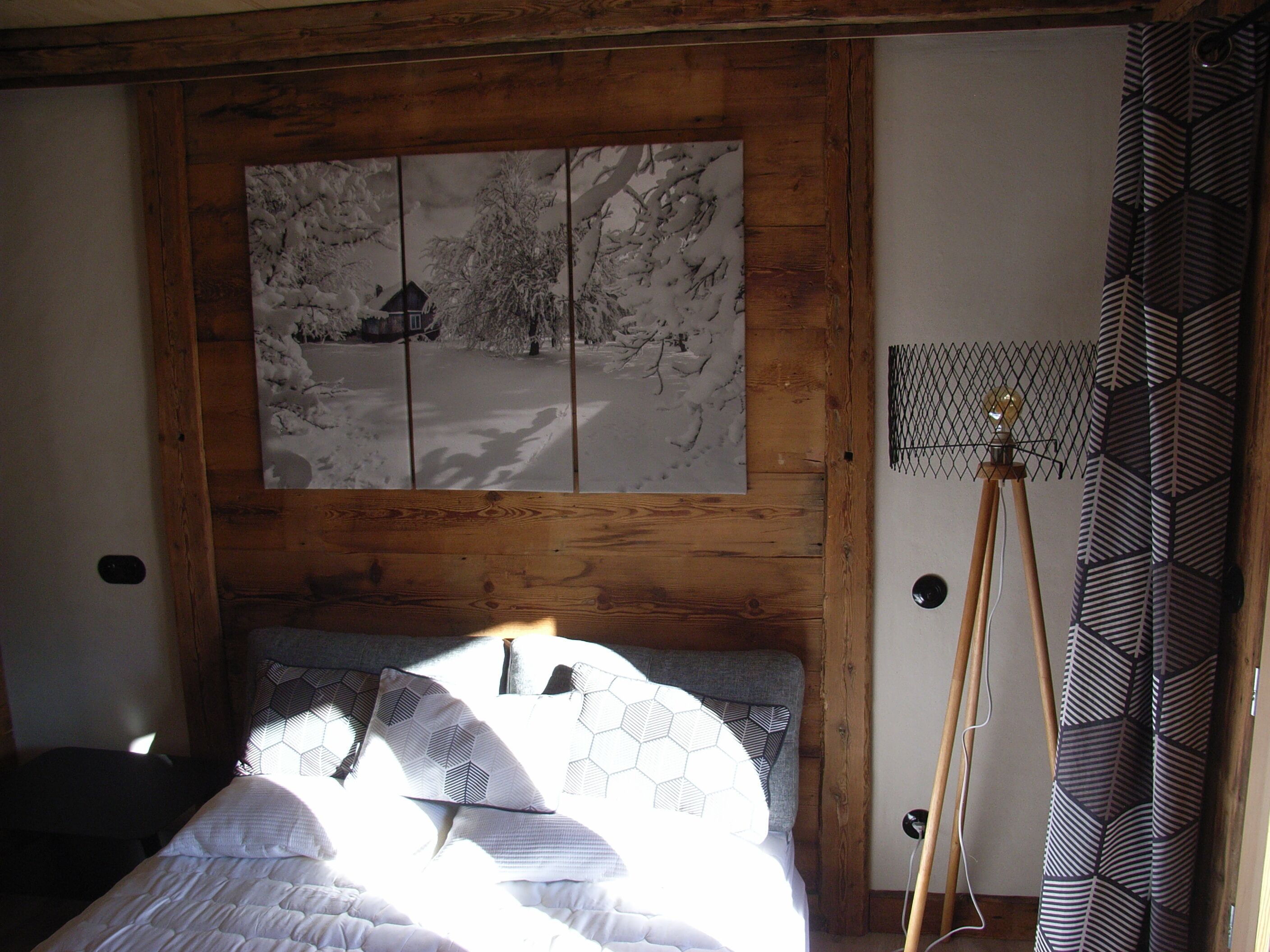 Un dormitorio con cabecera de madera, cama con ropa de cama blanca, un cuadro colgado en la pared de un paisaje nevado y una lámpara de pie.