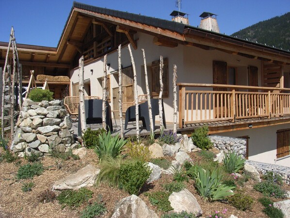Ein rustikales Chalet mit Holzbalkon, Steinmauern und einem Garten mit Steinen und Pflanzen.
