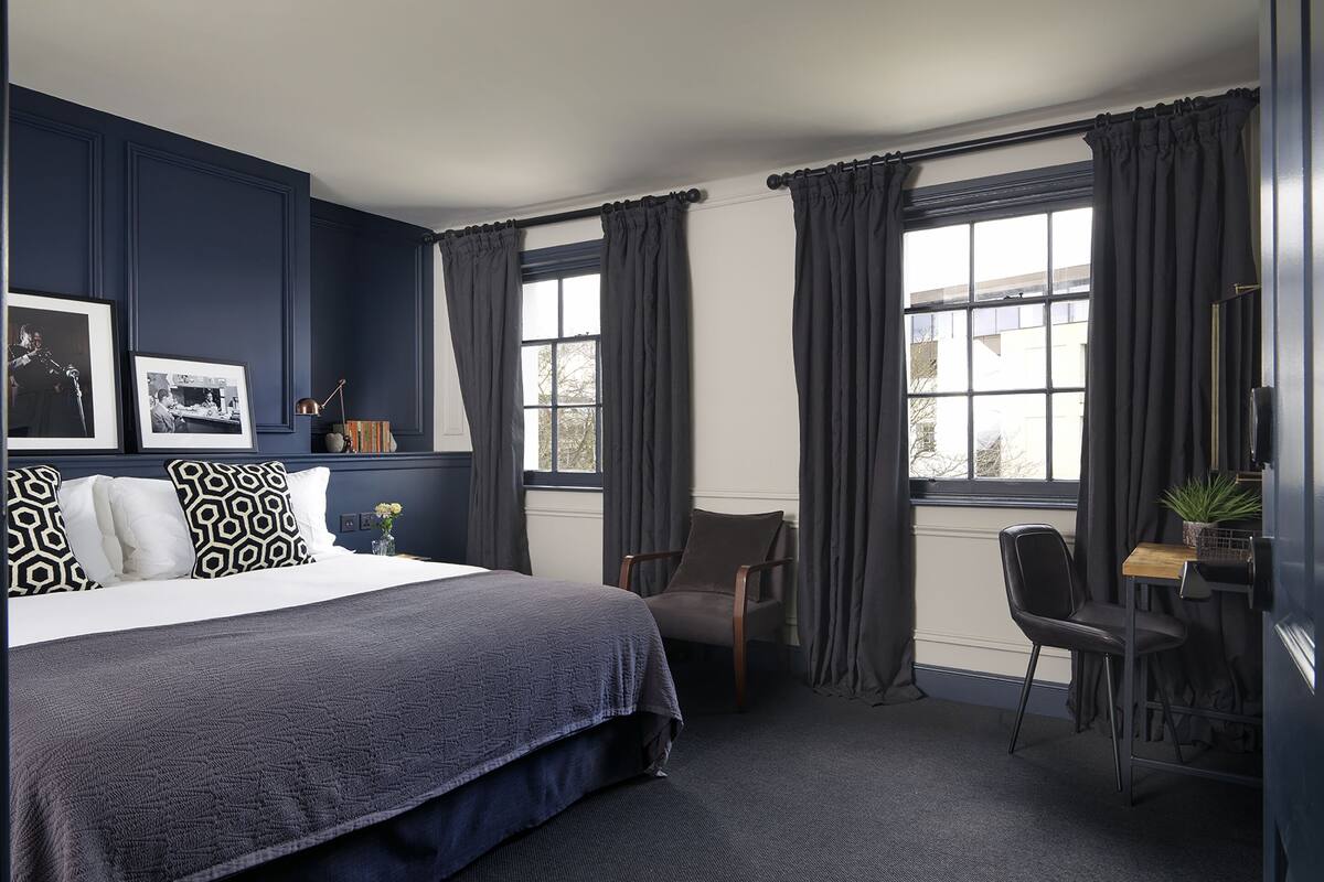 A hotel room with a large bed, a desk, and two chairs. There are curtains on the windows, and a plant is on the desk.