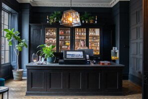 A bar area with a dark wood counter, a self-service check-in station, and shelves stocked with bottles. - The George (Cheltenham)