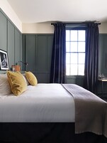 Una cama bien hecha con ropa blanca y dos almohadas amarillentas, un cuadro en la pared y una ventana con cortinas oscuras.