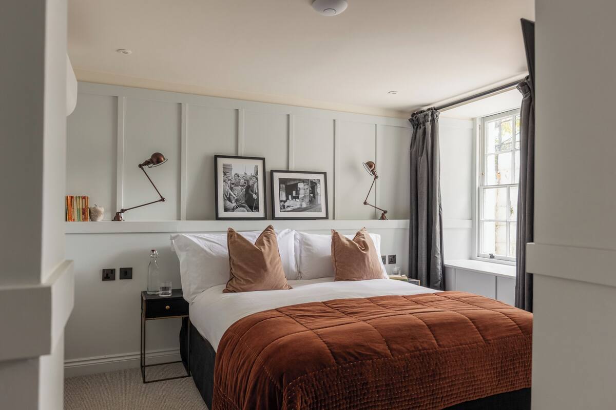 A bedroom with a large bed, a nightstand, a lamp, framed pictures on the wall, and a window with curtains.