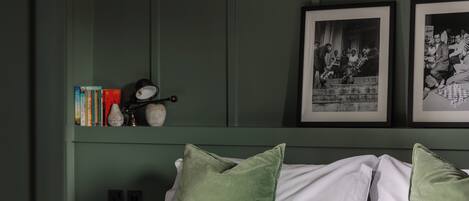 A neatly made bed with a quilted cover, green pillows, and a shelf with books and a magnifying glass.